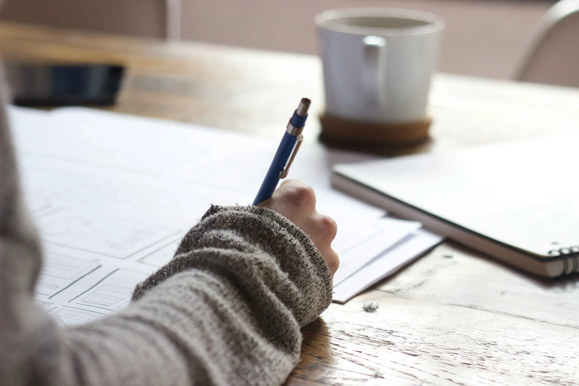 Ein Bild des Arms einer Person, die mit einem Stift auf Papier schreibt, mit einem Notizbuch und einer Kaffeetasse in der Nähe als symbolisches Bild für die Kategorie Bildung und Beratung.